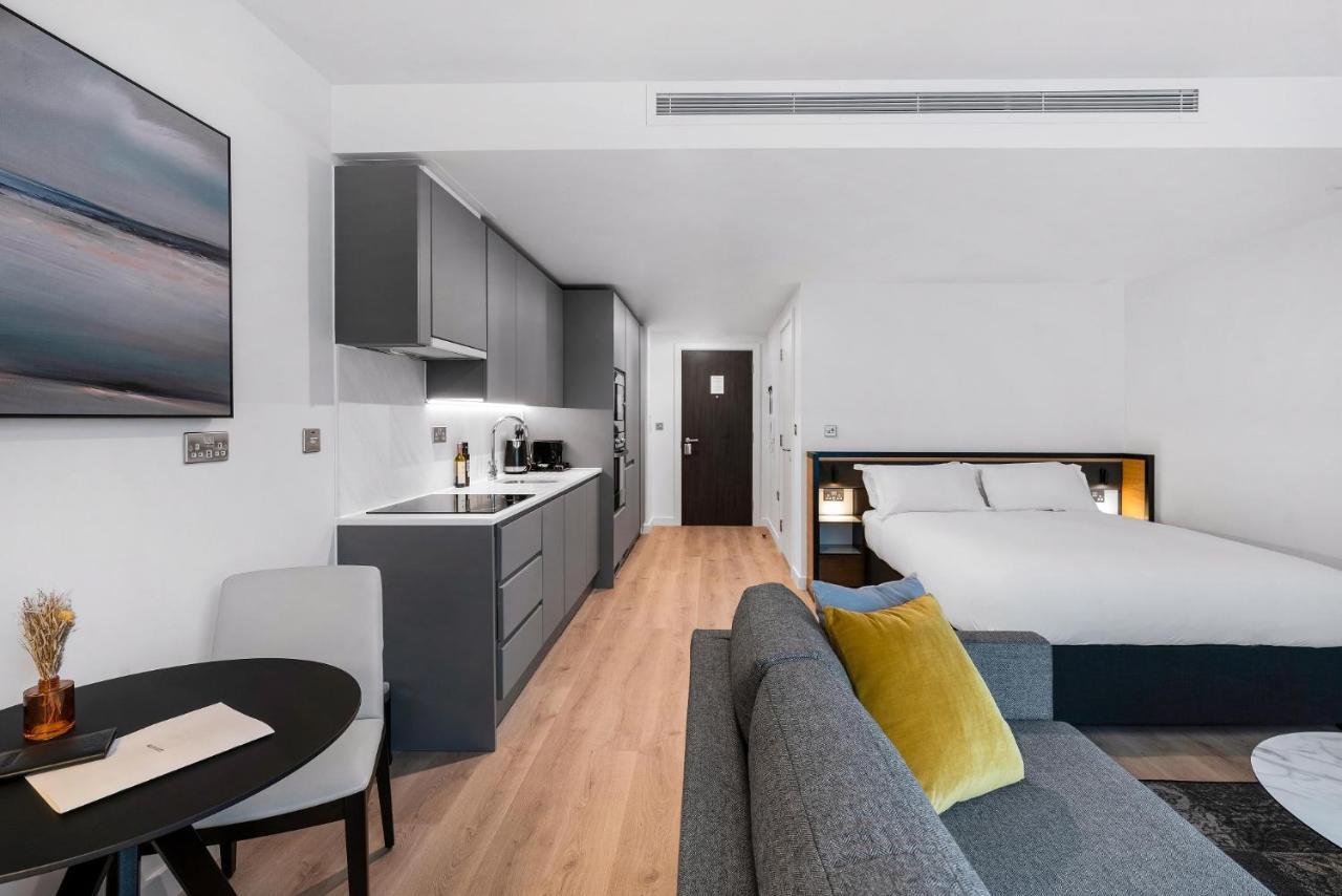 Citysuites 2 Aparthotel Manchester Zewnętrze zdjęcie The photo shows a modern and stylish studio apartment. It features a kitchenette with dark cabinetry and a small dining table with two chairs. There's a cozy sofa with colorful cushions, positioned next to the table. In the background, a large bed wi