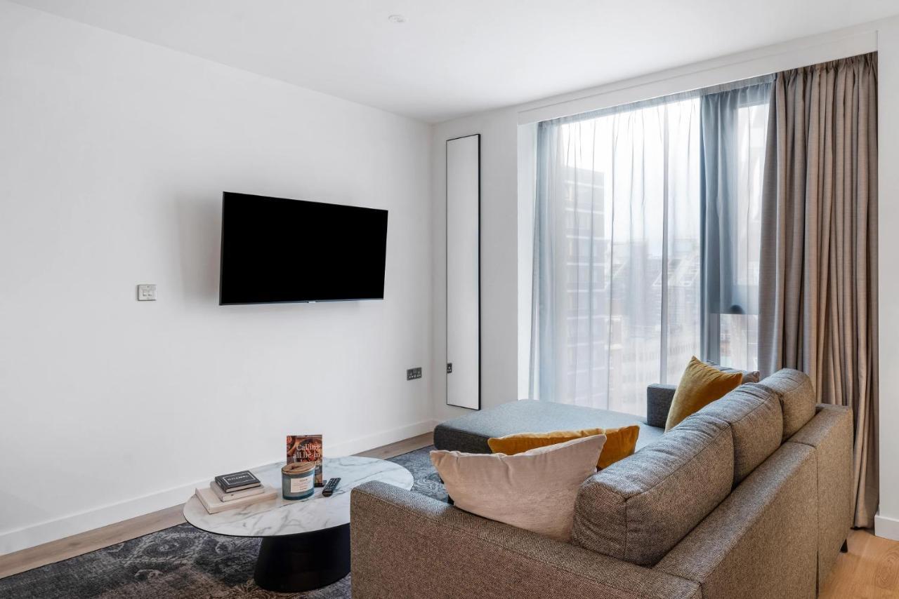 Citysuites 2 Aparthotel Manchester Zewnętrze zdjęcie The image shows a modern living room with a minimalist design. There is a large flat-screen TV mounted on the white wall. A comfortable gray sofa with textured cushions occupies the center of the room, complemented by a round coffee table in front of