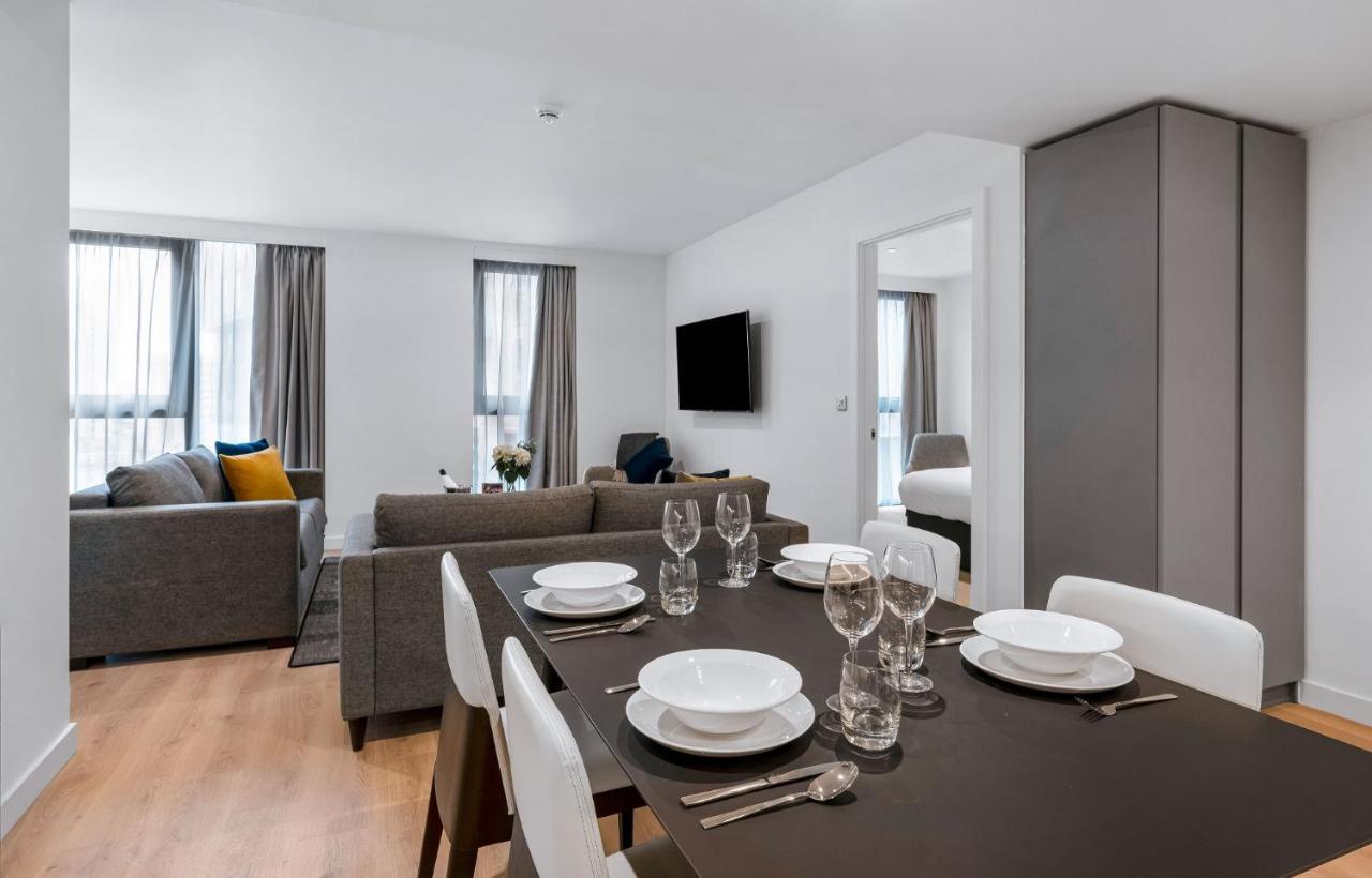 Citysuites 2 Aparthotel Manchester Zewnętrze zdjęcie The photo shows a modern living space with an open layout. In the foreground, there is a dining table set for a meal, featuring multiple plates, glasses, and cutlery arranged neatly. In the background, there is a comfortable-looking gray sofa with a 