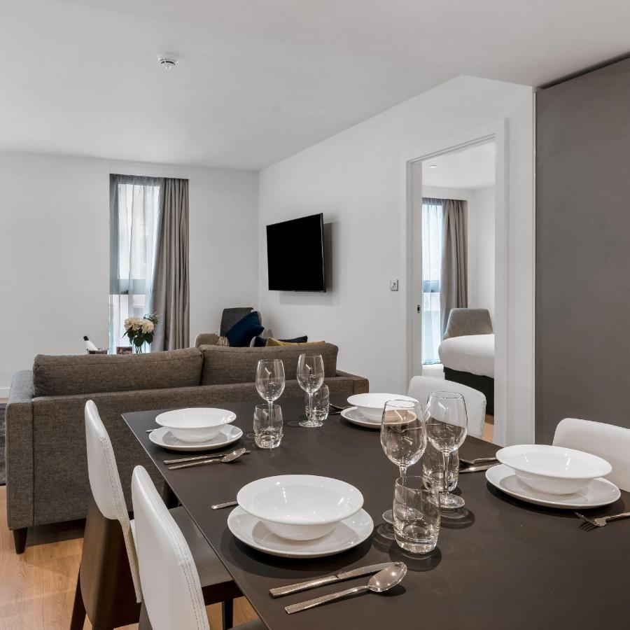 Citysuites 2 Aparthotel Manchester Zewnętrze zdjęcie The photo shows a modern, well-lit living space that features a dining area and a sitting area. In the foreground, there is a dining table set for six, with white plates and crystal glasses arranged neatly. The table is surrounded by white chairs. 

