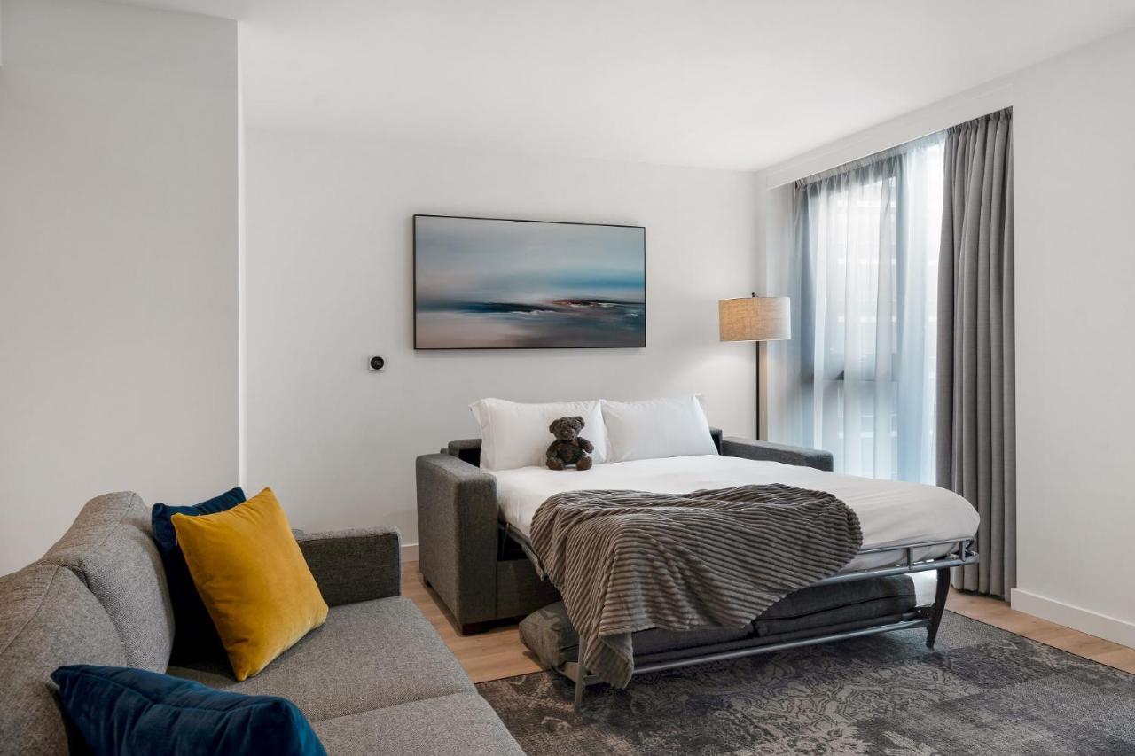 Citysuites 2 Aparthotel Manchester Zewnętrze zdjęcie The photo shows a modern and minimalistic guest room. There is a sofa on the left side, adorned with a colorful cushion. In the middle is a bed that appears to be a pull-out style, featuring white bedding and a gray throw blanket. A stuffed toy is pl