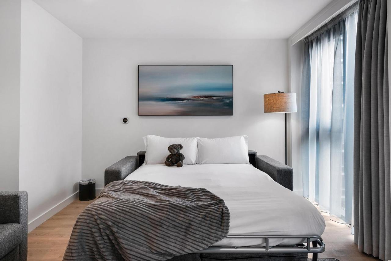 Citysuites 2 Aparthotel Manchester Zewnętrze zdjęcie The photo shows a modern bedroom featuring a bed with white bedding and a decorative throw blanket. There is a teddy bear placed on the bed. A piece of abstract art is hanging on the wall above the bed. The room has a large window with sheer curtains