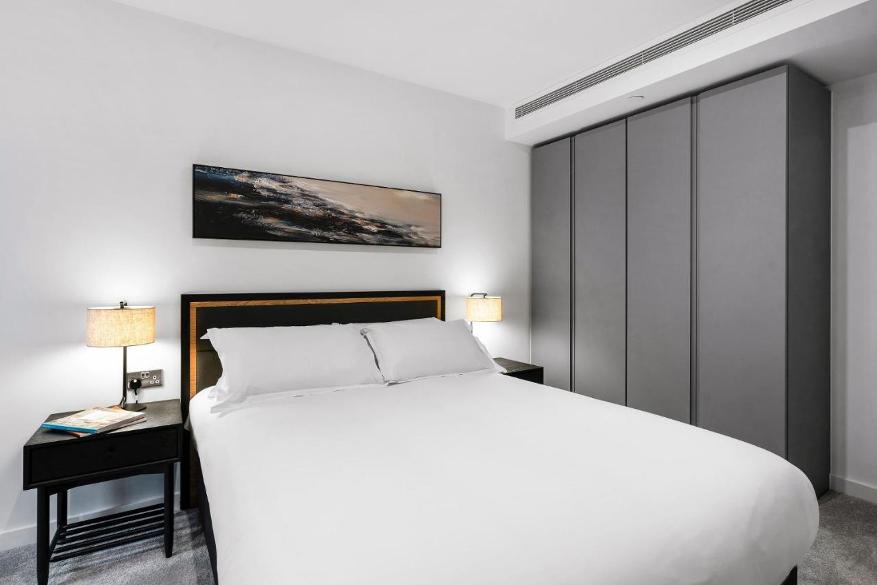 Citysuites 2 Aparthotel Manchester Zewnętrze zdjęcie The photo shows a modern bedroom designed with a minimalist aesthetic. The focal point is a large bed with white bedding, positioned against a wall. On either side of the bed, there are black nightstands topped with lamps, providing a warm glow. Abov