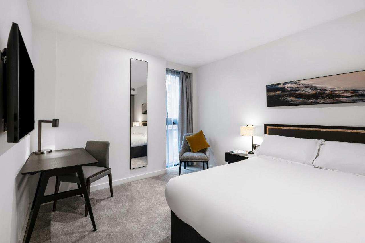 Citysuites 2 Aparthotel Manchester Zewnętrze zdjęcie The photo shows a modern hotel room with a clean and minimalist design. It features a large bed with white linens, a small desk with a chair, and a mirror mounted on the wall. There is a chair in one corner with a decorative pillow, and the room has 