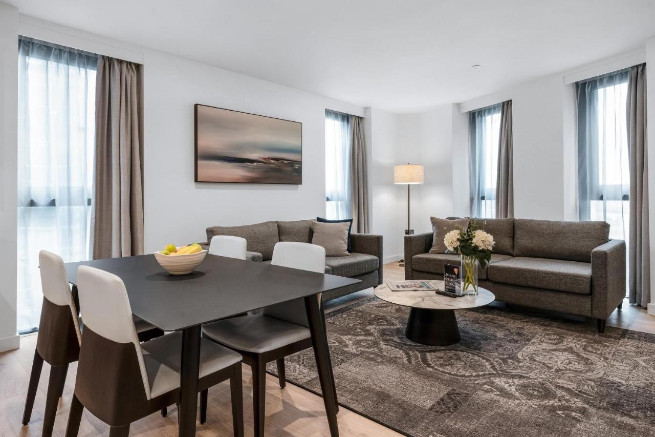 Citysuites 2 Aparthotel Manchester Zewnętrze zdjęcie The photo depicts a modern and stylish interior of a living space. On the left, there is a dining table with four chairs, featuring a bowl of fruits on the table. In the background, there's a cozy seating area with a gray sofa and a smaller round cof