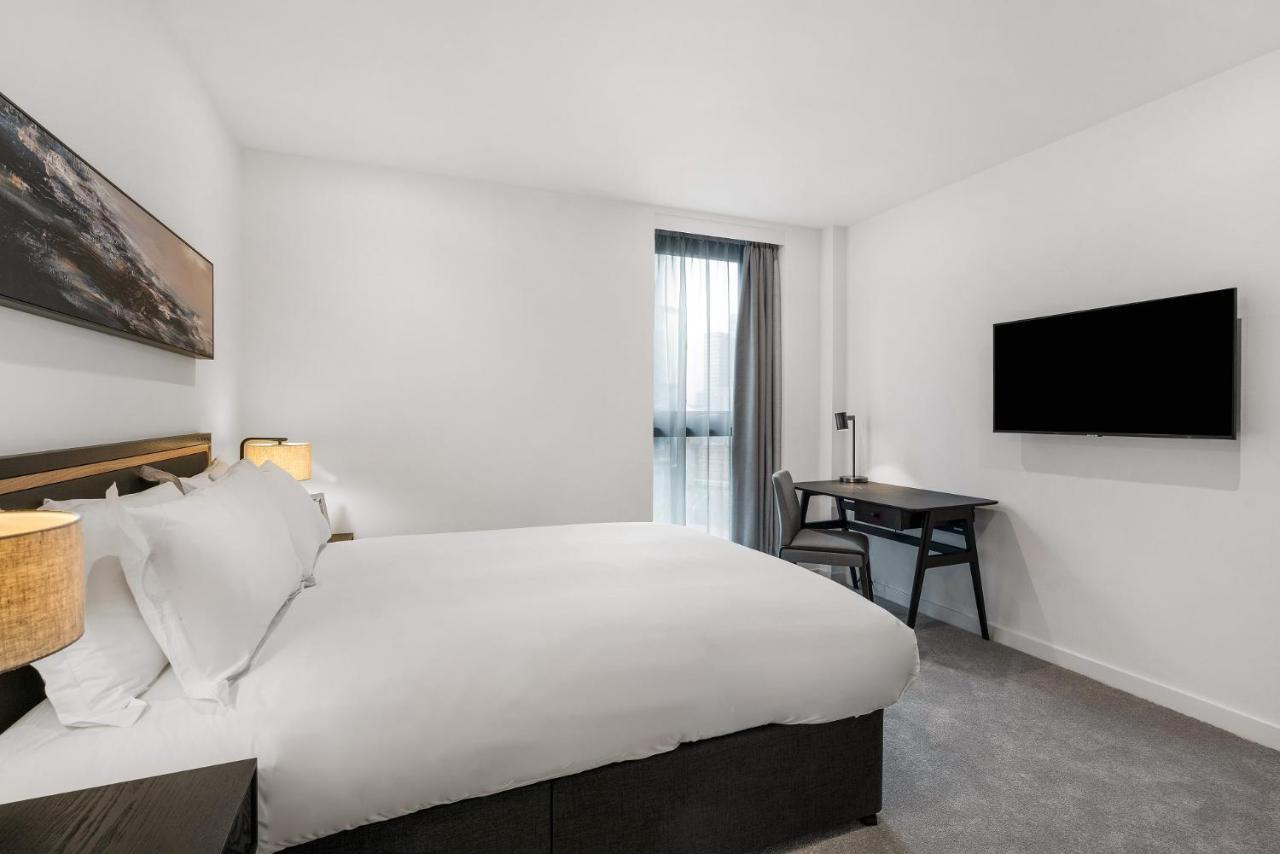 Citysuites 2 Aparthotel Manchester Zewnętrze zdjęcie The photo shows a modern hotel room featuring a large bed with white linens and a dark headboard. There are two bedside lamps on either side of the bed. A desk with a chair is placed against one wall, and a flat-screen TV is mounted on another wall. 