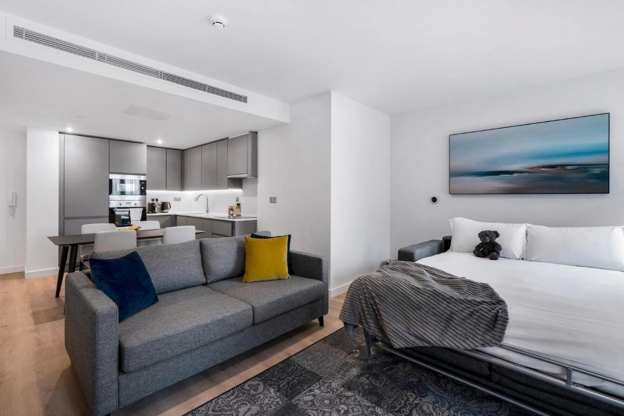 Citysuites 2 Aparthotel Manchester Zewnętrze zdjęcie The photo shows a modern, open-concept living space that includes a kitchen and a sleeping area. 

- **Living Area**: On the left, there's a gray sofa with a blue and yellow throw pillow. A blanket is draped over one side, and there's a small dark ru