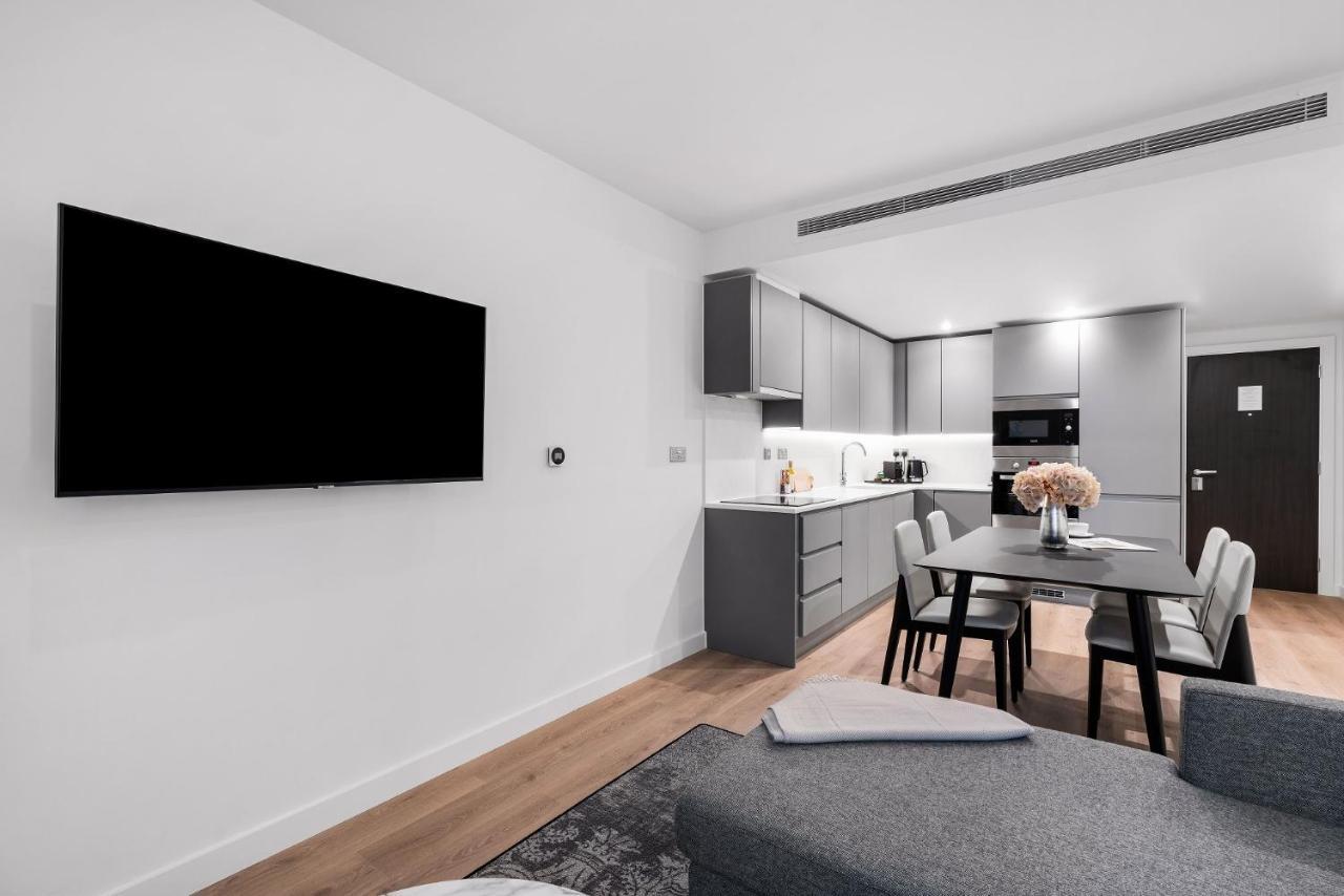 Citysuites 2 Aparthotel Manchester Zewnętrze zdjęcie The photo shows a modern, minimalist living space. On the left, there is a large flat-screen TV mounted on the wall. To the right, a kitchen area features sleek gray cabinets and appliances, including a built-in microwave. In front of the kitchen, th