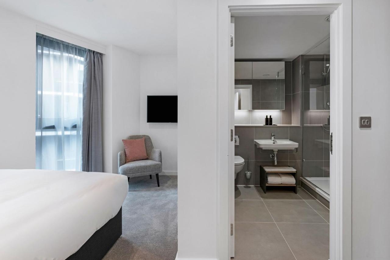 Citysuites 2 Aparthotel Manchester Zewnętrze zdjęcie The photo shows a modern hotel room and bathroom setup. On the left, there is a bed with white bedding and a small sitting area that includes a gray chair and a pillow in a subtle pink shade. A flat-screen TV is mounted on the wall, providing enterta