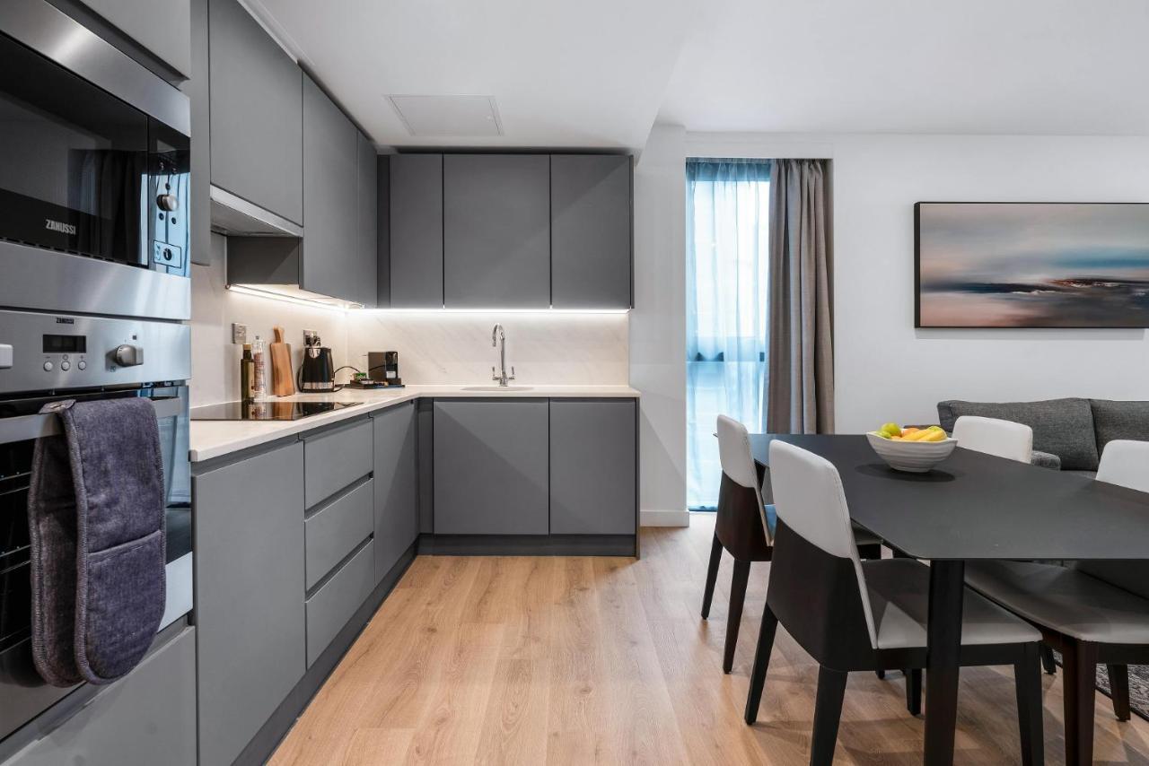 Citysuites 2 Aparthotel Manchester Zewnętrze zdjęcie The photo shows a modern kitchen and dining area. The kitchen features sleek gray cabinetry, a white countertop, and built-in appliances such as an oven. There is a sink and some kitchen utensils displayed. The adjacent dining area contains a black t