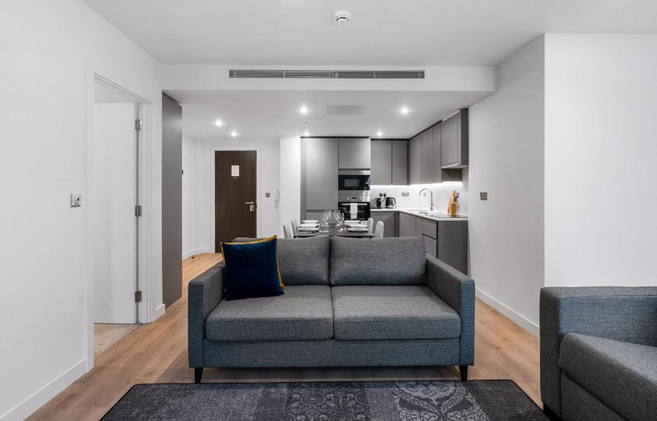 Citysuites 2 Aparthotel Manchester Zewnętrze zdjęcie The photo shows a modern interior space, likely a living area or apartment. In the foreground, there is a gray sofa with a blue cushion positioned on a textured rug. To the back, there is an open kitchen with sleek cabinetry and appliances, visible a