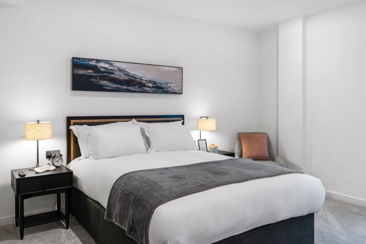 Citysuites 2 Aparthotel Manchester Zewnętrze zdjęcie The photo shows a modern bedroom featuring a large bed with crisp white linens and a grey throw blanket. On either side of the bed, there are bedside tables with lamps. A piece of abstract art hangs above the headboard, and there is a chair in a corn