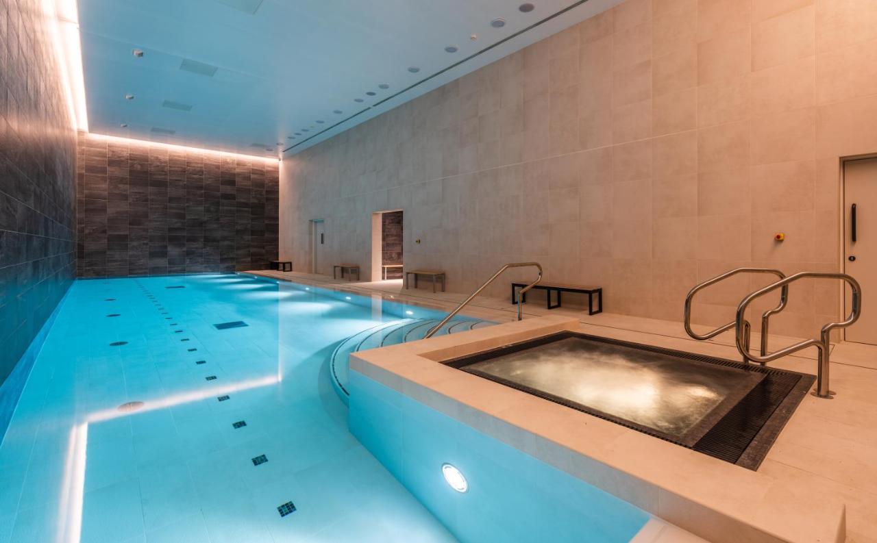 Citysuites 2 Aparthotel Manchester Zewnętrze zdjęcie The photo shows an indoor swimming pool area. The pool is a bright blue color and features smooth, tiled surfaces. On one side, there is a hot tub area with steps leading into it. The walls are made of light-colored stone, and there is ambient lighti