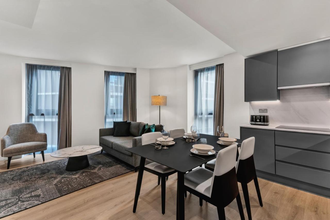 Citysuites 2 Aparthotel Manchester Zewnętrze zdjęcie The photo shows a modern living space that includes a dining area and a seating area. There's a sleek black dining table set with plates and cutlery, accompanied by white and black chairs. To one side, a comfortable gray sofa faces the dining area, c