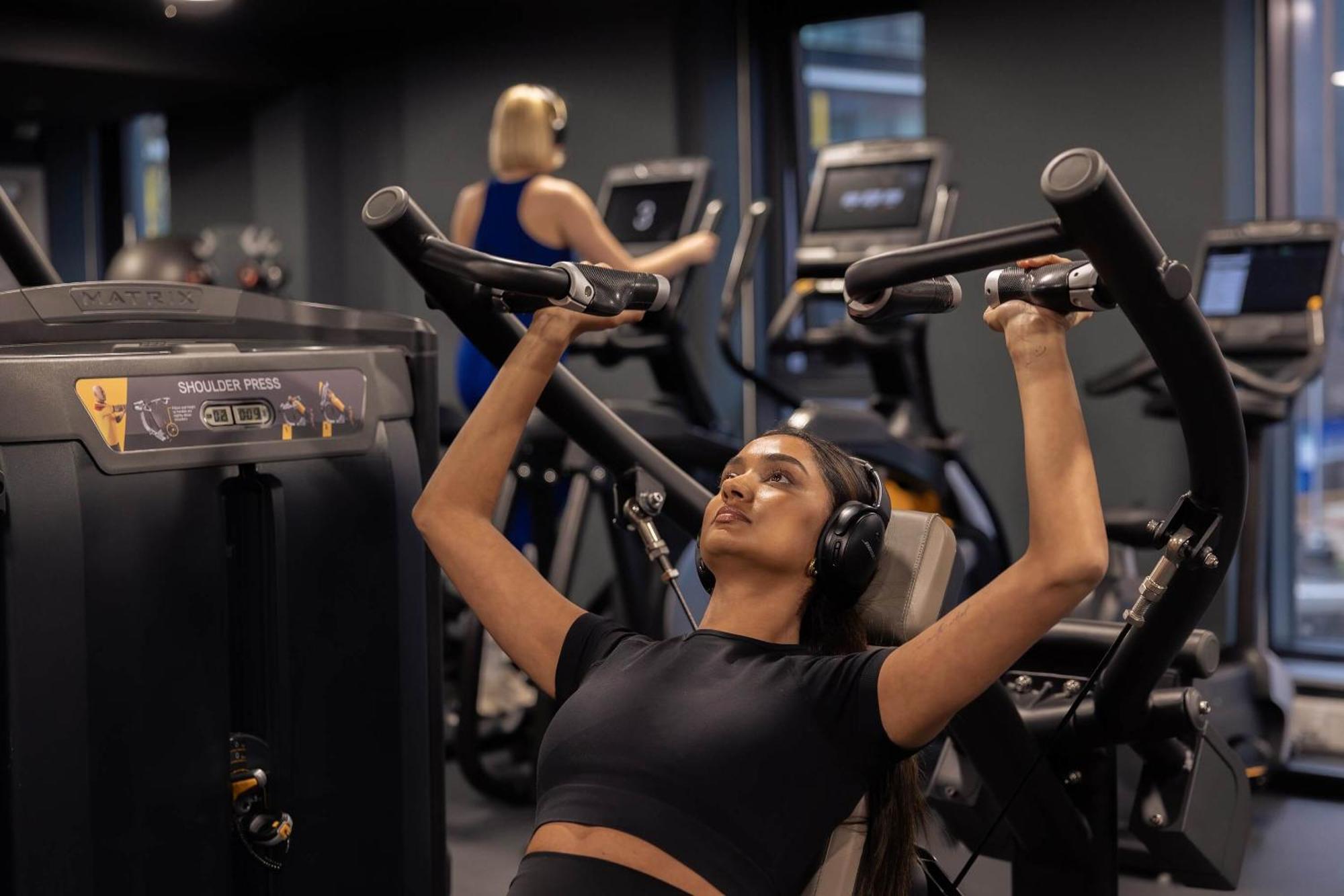 Citysuites 2 Aparthotel Manchester Zewnętrze zdjęcie The photo shows a gym setting with two individuals engaged in physical exercise. In the foreground, a woman is using a chest press machine while wearing headphones, focusing on her workout. She appears to be in a seated position, pushing the handles 