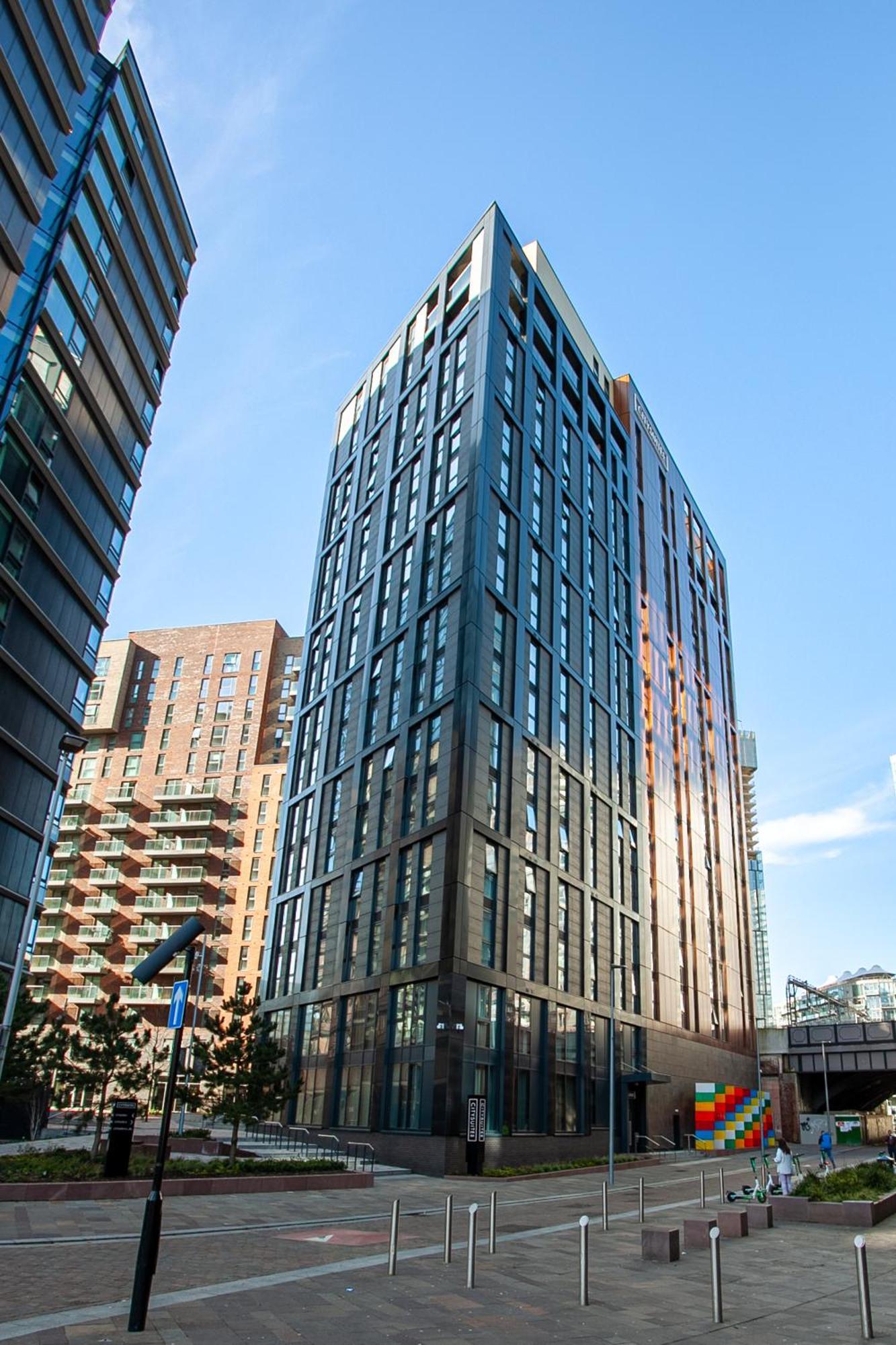 Citysuites 2 Aparthotel Manchester Zewnętrze zdjęcie The photo shows a modern high-rise building with a sleek, glass façade. It is set against a blue sky, indicating a clear day. The building appears to have multiple floors with large windows that reflect light. Nearby, there are additional buildings, 